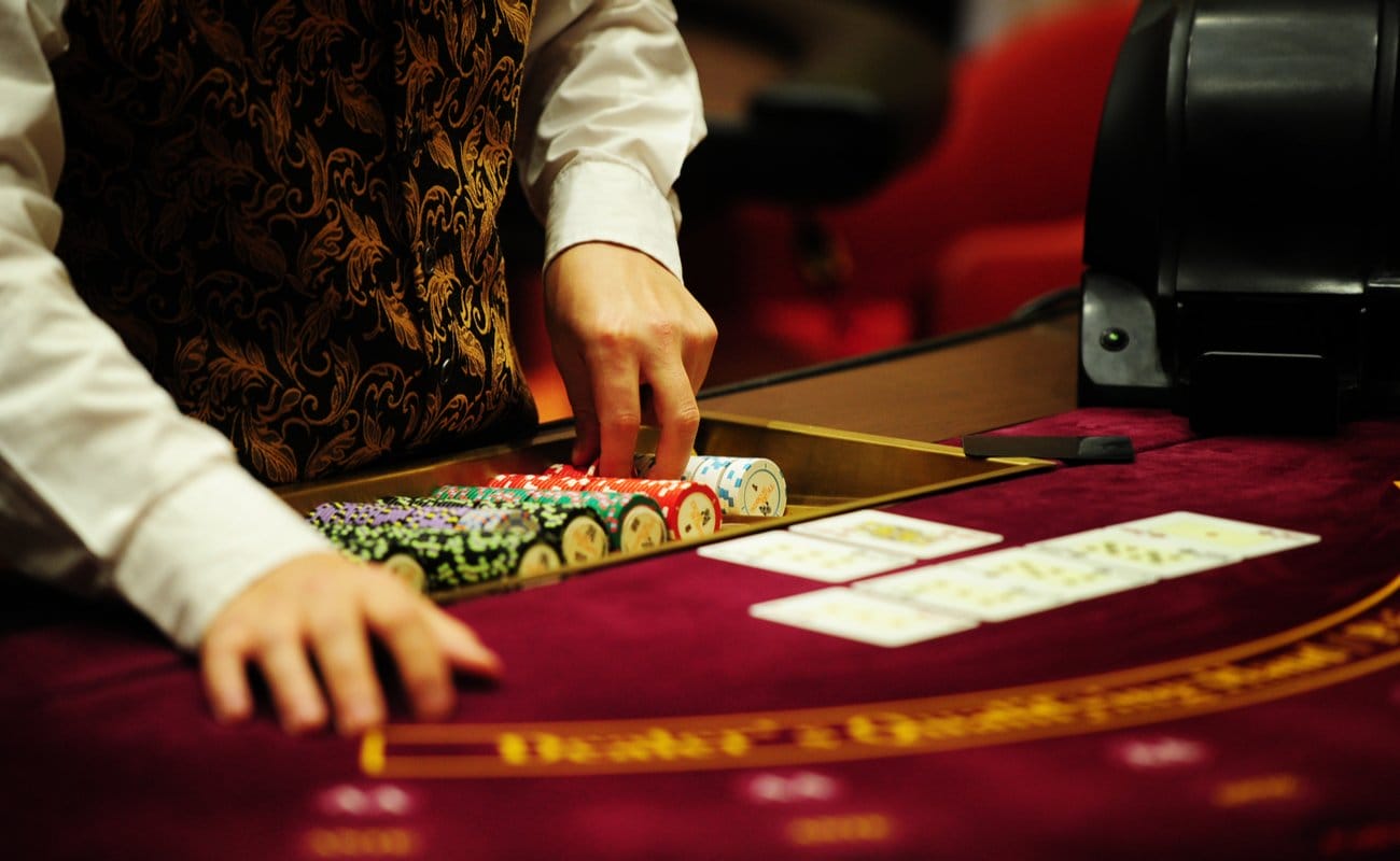 A dealer stands with chips, and playing cards at a red felt casino table.