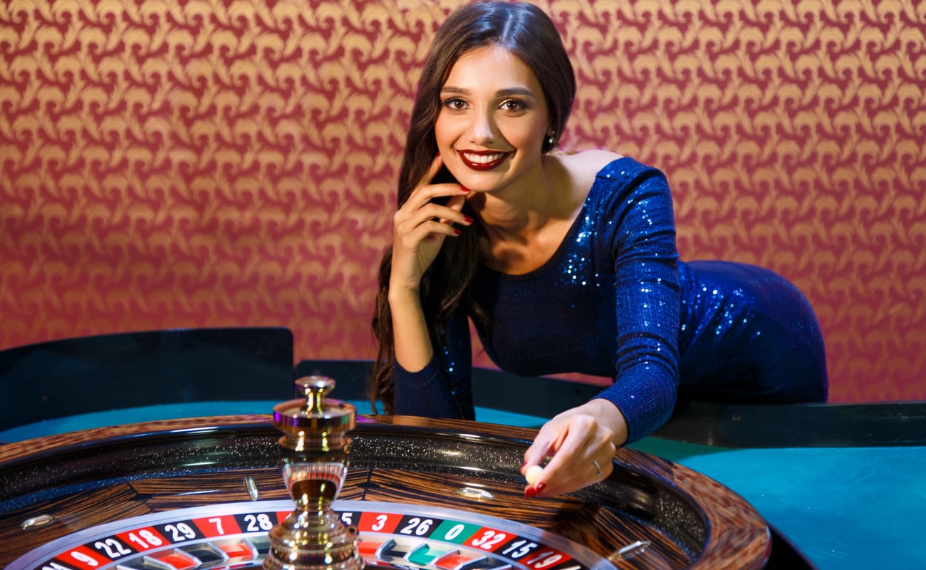 Female croupier in a blue dress leans over a roulette wheel on a blue felt casino table.