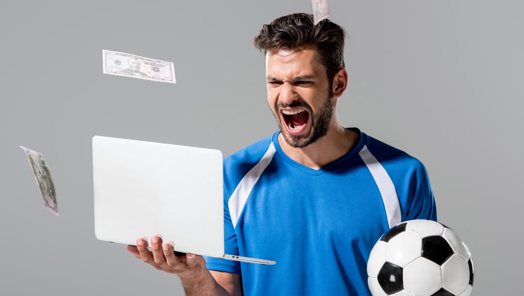 A football player in uniform screaming with excitement in front of a laptop while playing online slots and winning a big payout.