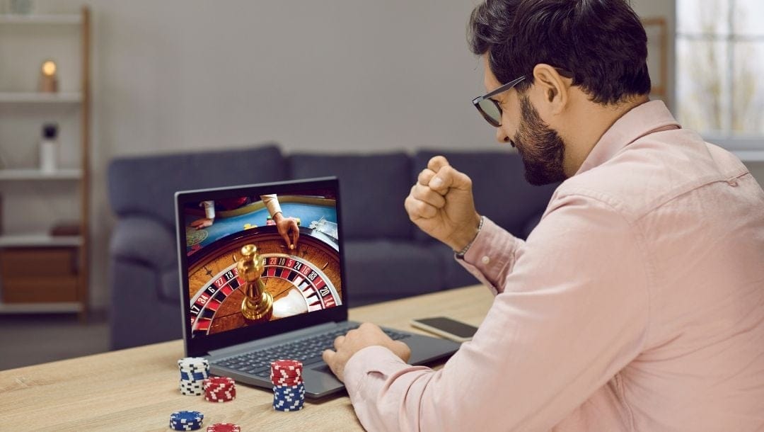 A person sits in front of their laptop and raises a fist in victory after winning an online live dealer roulette game.