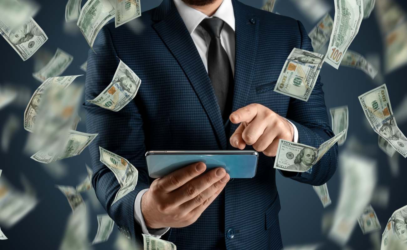 Man in a suit playing games on his mobile phone with 100-dollar bills floating around him