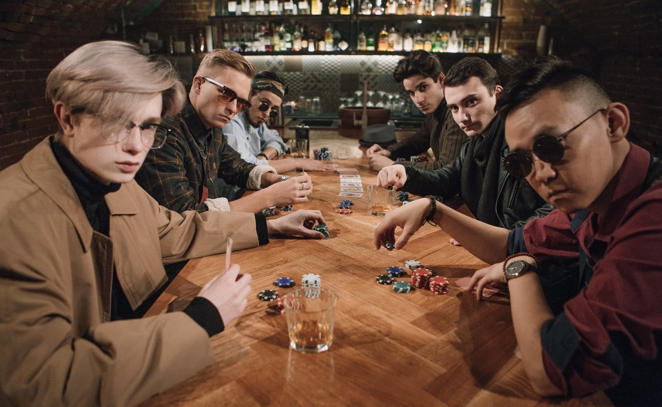 Six men playing poker at a wooden table with a bar in the background.