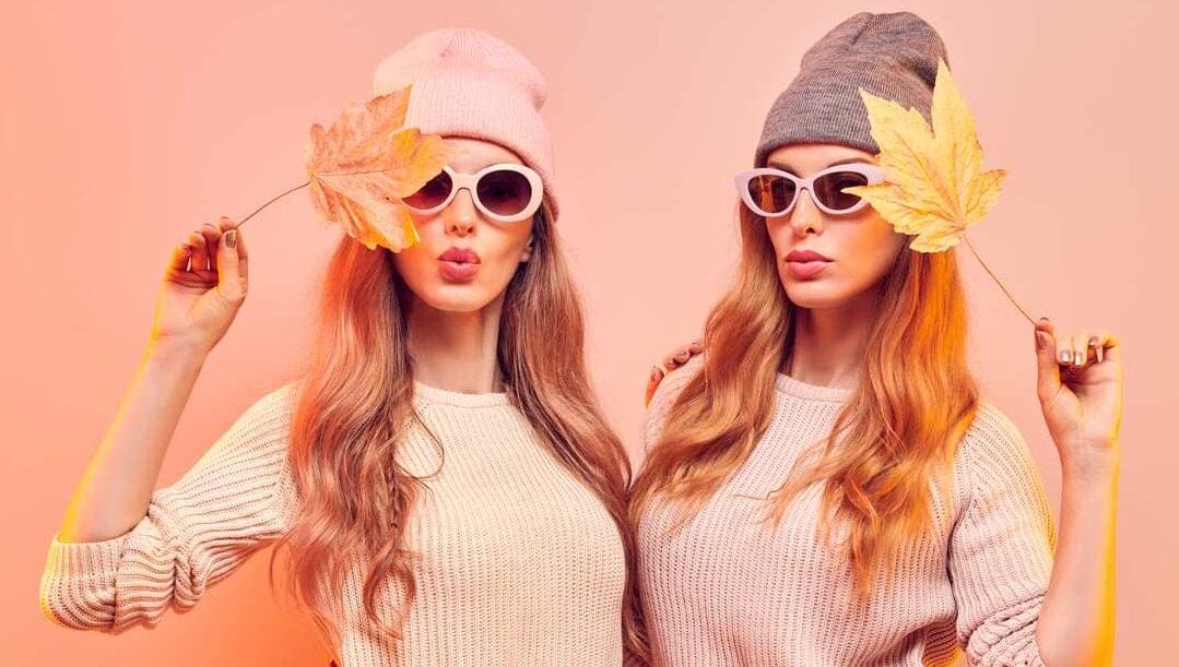 Two women against a pink background wearing beanies and sunglasses and holding fall leaves against their faces.