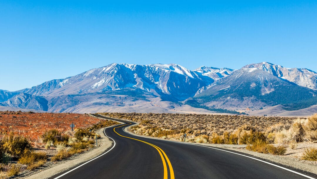 A road going through dry land with snow-capped mountains in the background.