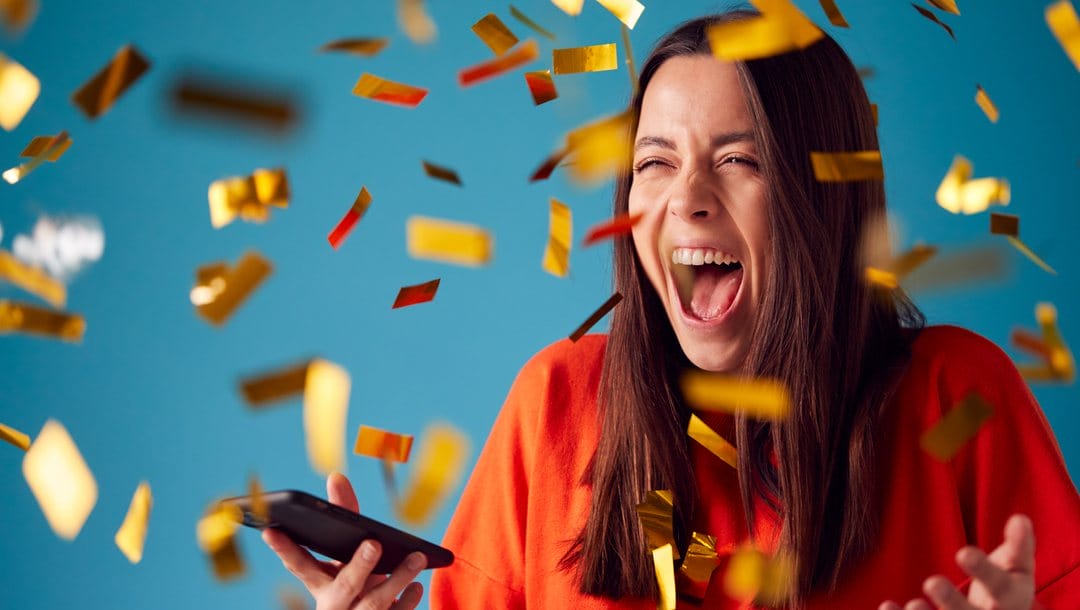 Woman holding a mobile phone shouts excitedly while being showered with gold confetti, against a blue background.