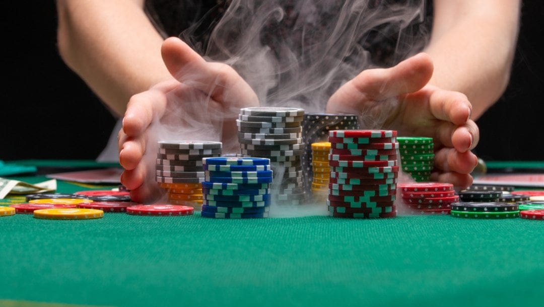 A woman cups her hands around stacks of poker chips on a casino table while smoke rises from beneath them.