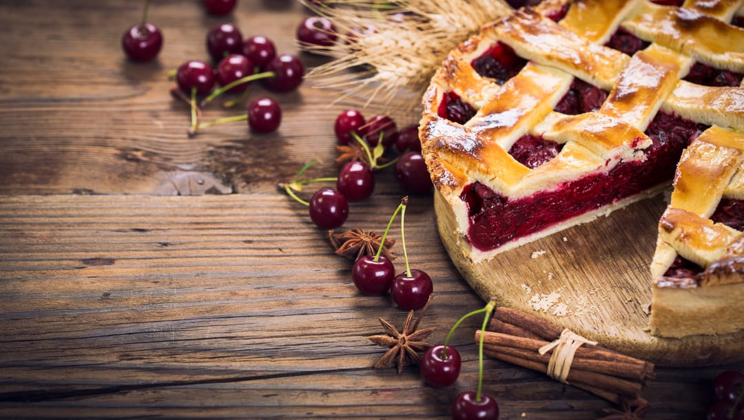 A cherry pie with a slice taken out on a table with cherries, star anise and cinnamon.