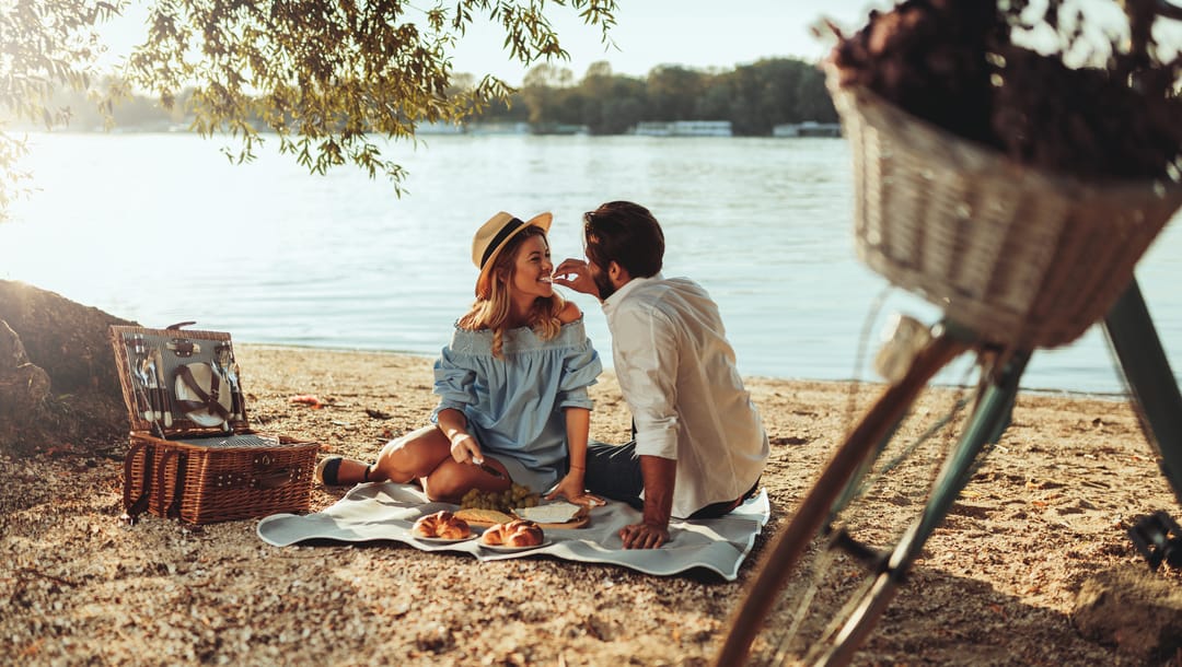 A man feeds a woman at a romantic picnic on the lakeside.