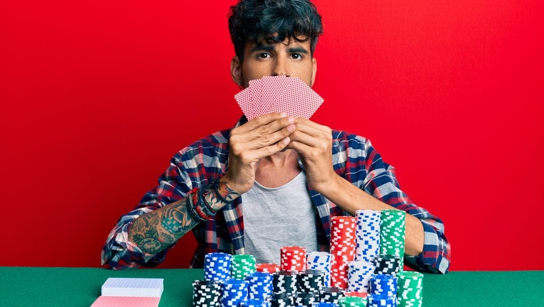 Young man playing poker and covering face with cards against a red background.