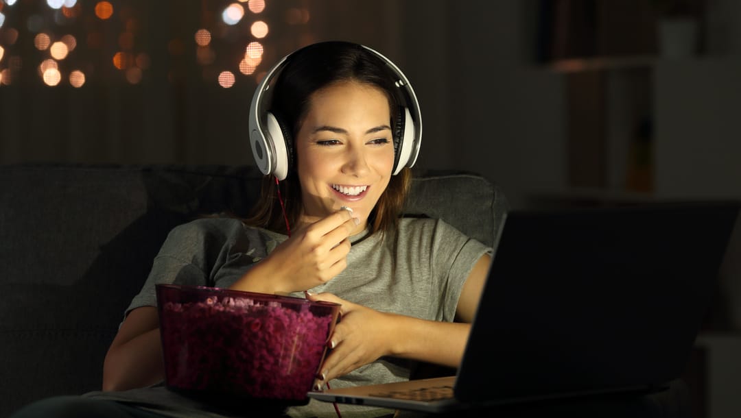 A woman wearing headphones eats popcorn at her laptop.