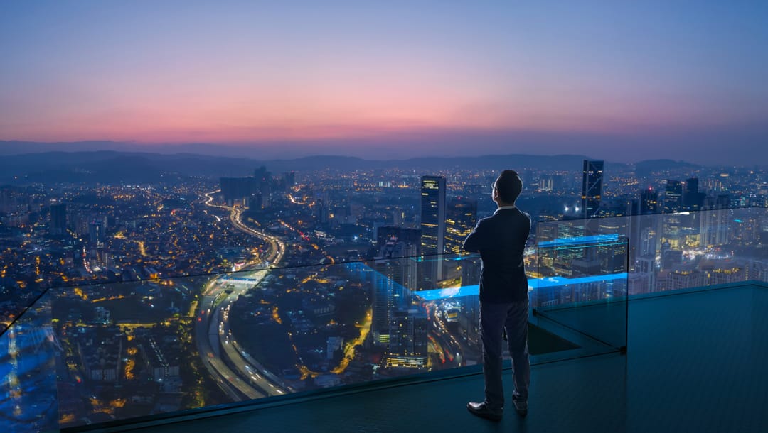 A businessman stands on an open rooftop and looks out at the cityscape as evening falls.