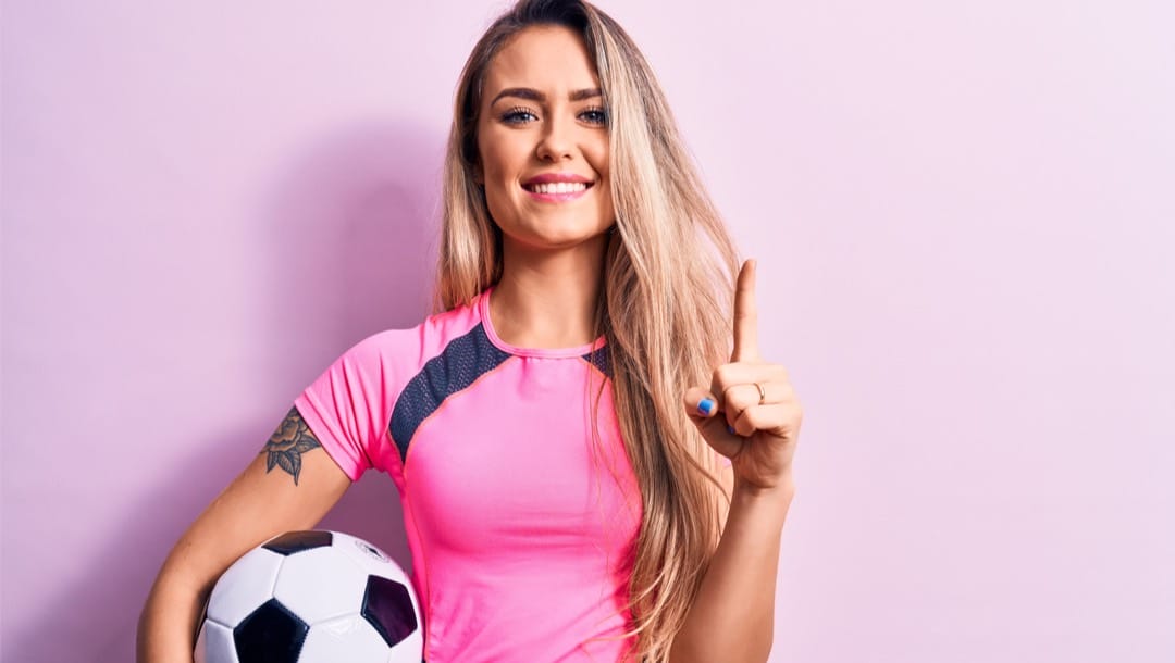 A blonde woman wearing a pink shirt holding a black and white soccer ball.