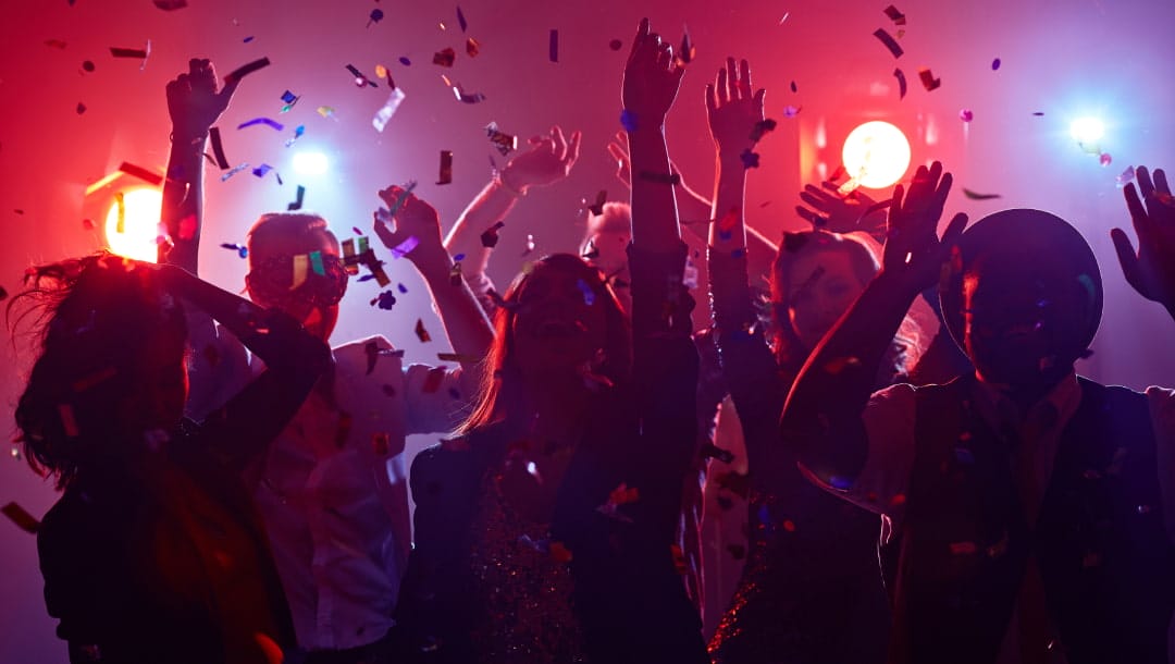 People with their hands up dancing to music in a nightclub as confetti rains down from the ceiling.