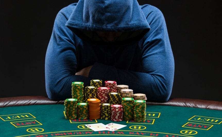 A poker player wearing a hoodie leans over his stacks of poker chips and his poker hand.