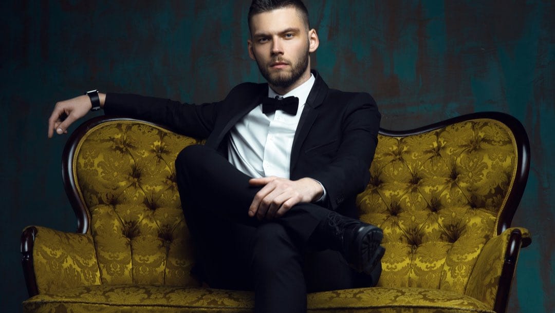 A man wearing a black tuxedo sitting on a gold couch.