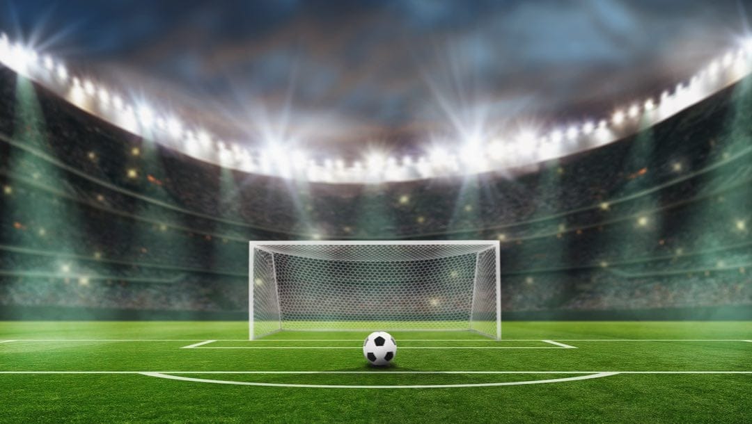 A soccer ball in front of the goal post in a large soccer stadium.