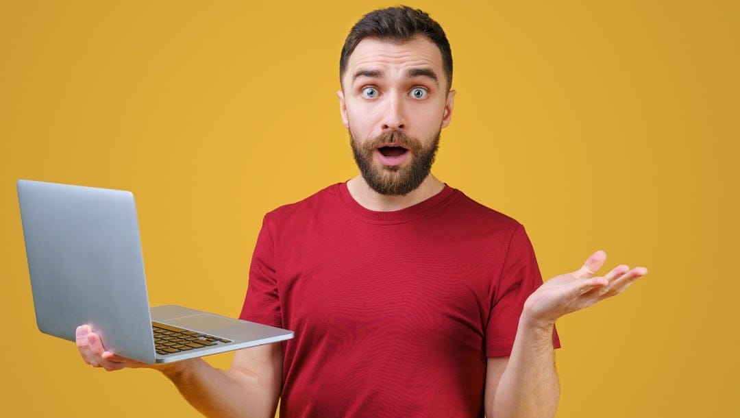 A confused bearded man holds an open laptop in one hand.