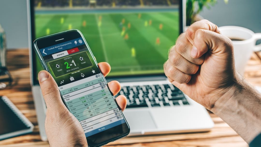 A man holding a smartphone featuring a sports betting app with a soccer game playing on his laptop.