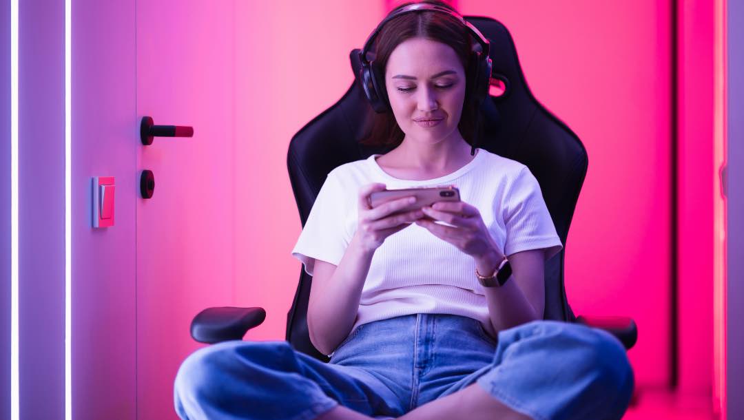 A woman sitting on a gaming chair with a headset and playing on a mobile phone.