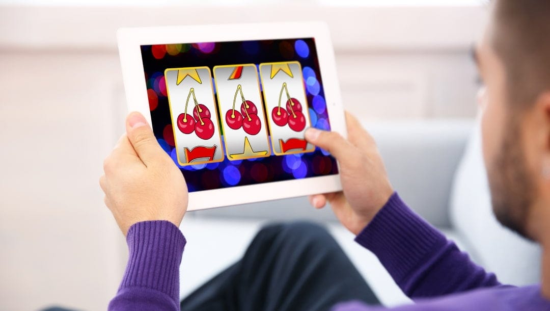 A person in a purple sweater playing a slot machine game on a white tablet. The screen shows a winning combination of cherries across all three reels, with a colourful, blurred background suggesting an online casino game interface.