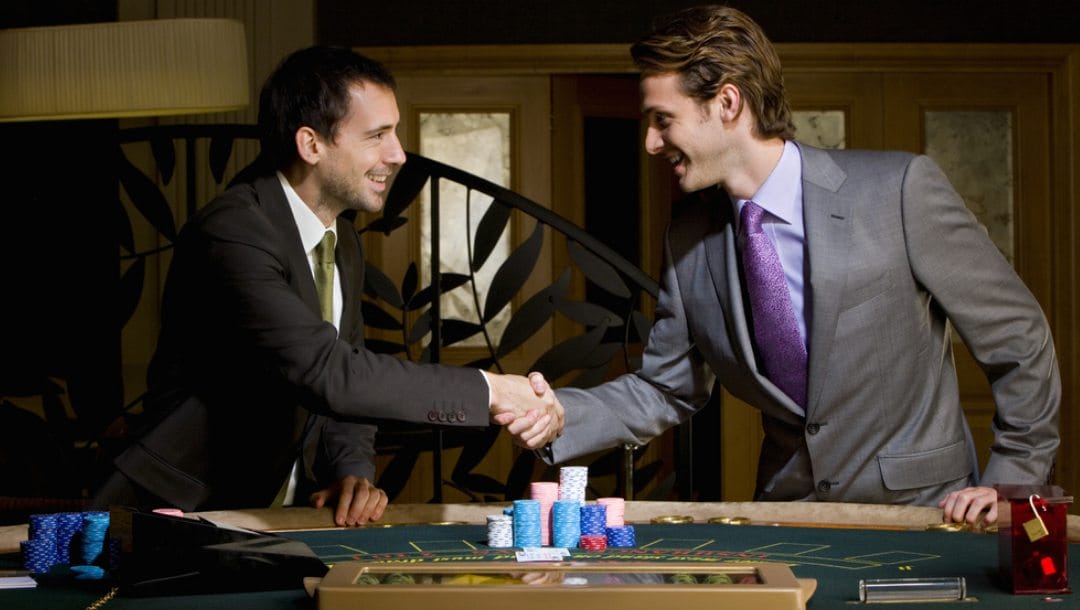 Two men in business suits shake hands next to a casino table.