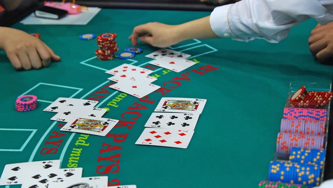 A blackjack table with a game in progress with hands and bets on the table. A dealer is reaching towards a player’s chips. There are four hands and the dealer’s hand visible on the table. There is a chip rack with casino chips in front of the dealer.