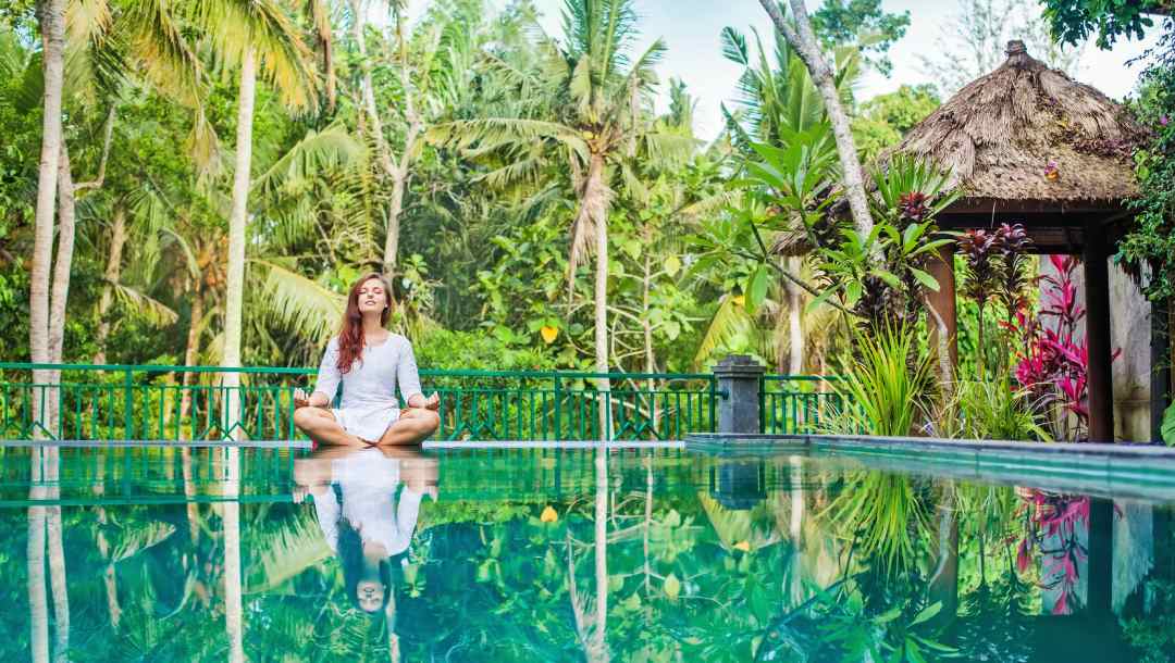 A woman meditating at a gorgeous poolside with tropical trees in the background.
