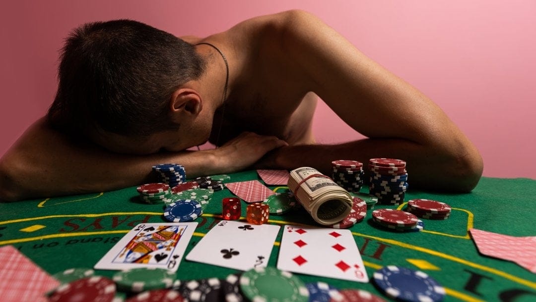 An upset poker player places his head on his arms on a poker table with cards, chips and dice.