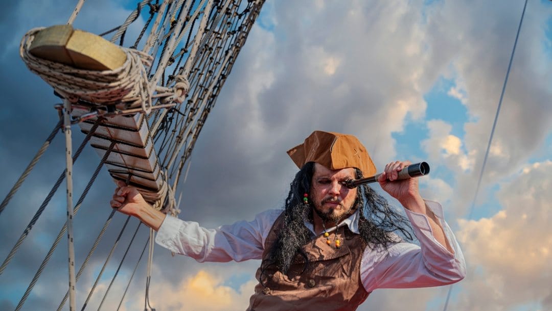 A man dressed as a pirate looking through a telescope from a ship.
