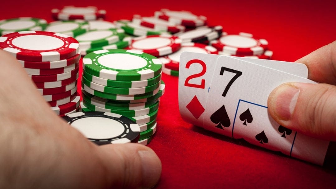 A poker player shows a hand with 2 and 7 and holds some poker chips on a poker table.