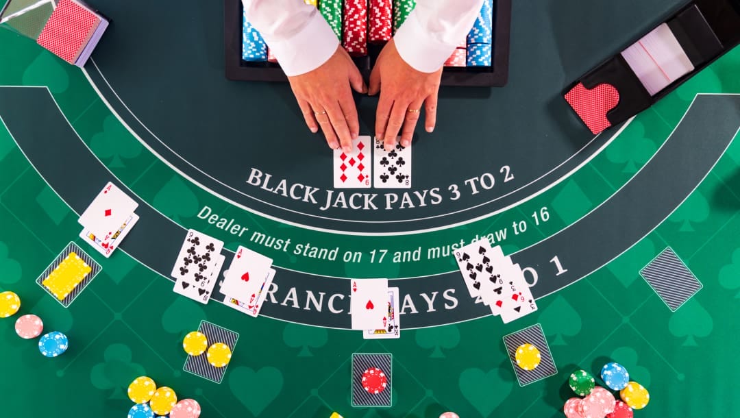 A top-down view of a green blackjack table. Five hands have been dealt and bet on. There are stacks of casino chips behind each of the bets. Both of the dealer’s cards are visible with one hand placed on each end of their cards. The card shoe is visible on the right of the dealer and stacks of playing cards on the left of the dealer.