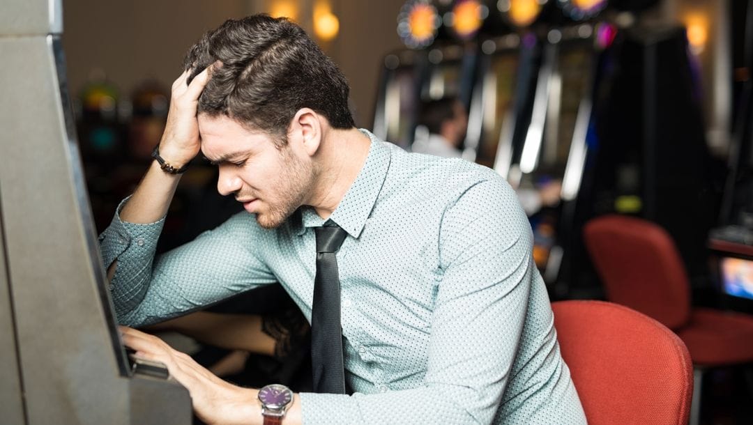 A frustrated man leans against a slot machine with his head in his hand.