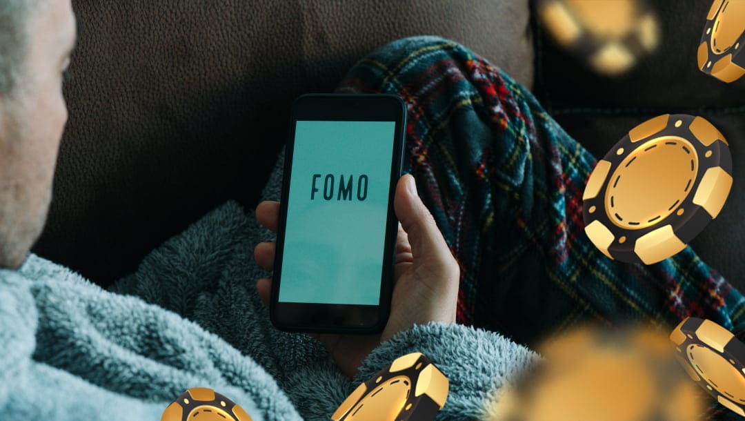 A man sitting on a brown couch in a gown and pyjama pants. The man is holding a smartphone with the word FOMO against a blue screen. There are digital black and gold casino chips floating around the screen.