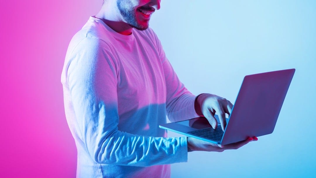A man standing and using a laptop while bathed in lavender and blue light.