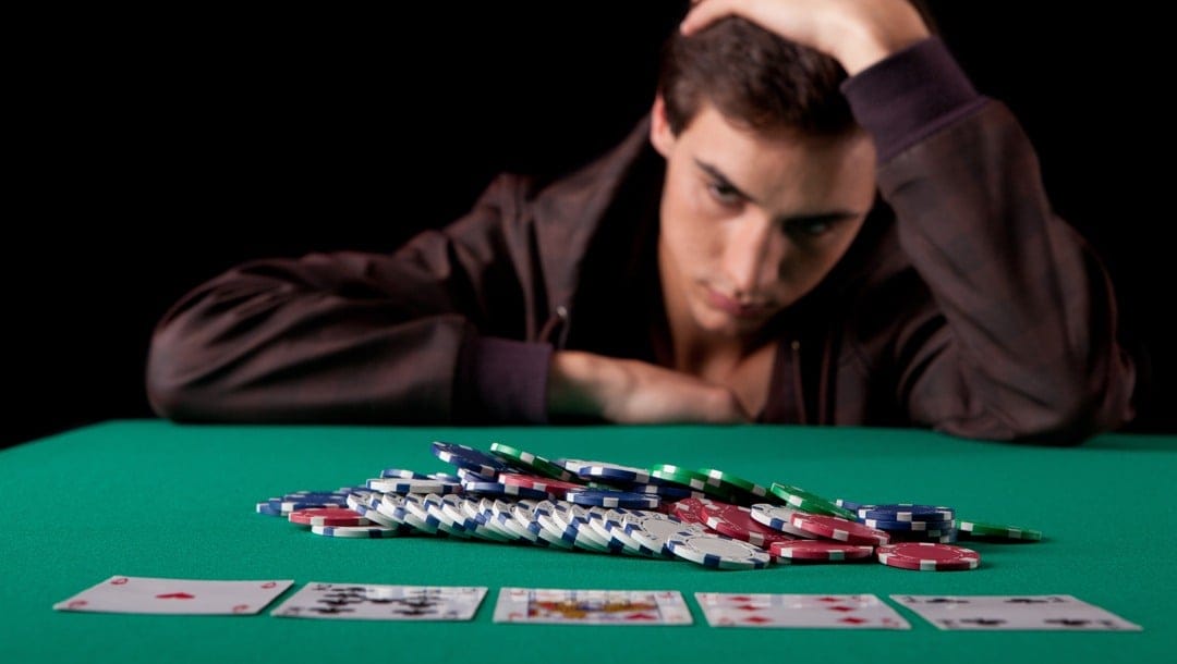 A despondent man with his hand on his head looking at poker chips and cards in the center of a table.