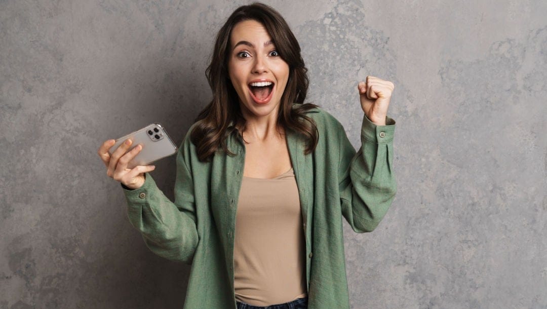 A woman making a winning gesture with one hand and holding a smartphone in the other.