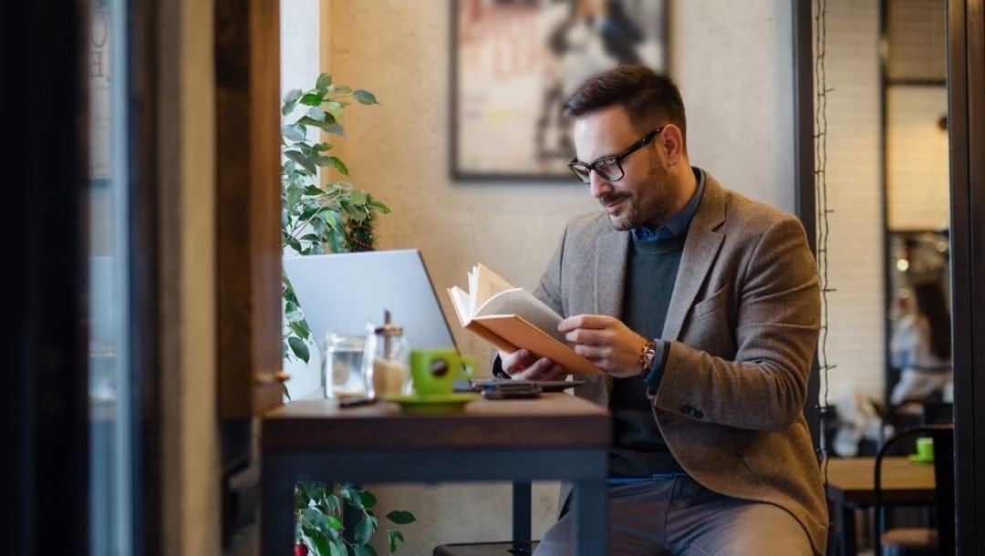 A smartly dressed man reading a book at a coffee shop with his laptop on the table.