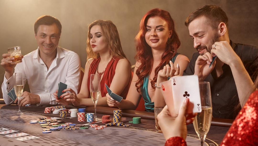 A group of well-dressed men and women playing poker and enjoying drinks at a poker table.