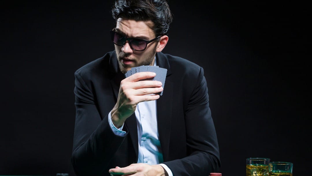 A person wearing a suit and sunglasses holds their poker hand up while looking to their left.