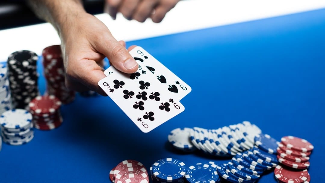 A poker player reveals two hole cards: a pair of nines. There are various stacks of chips in the background and the poker pot in the foreground.