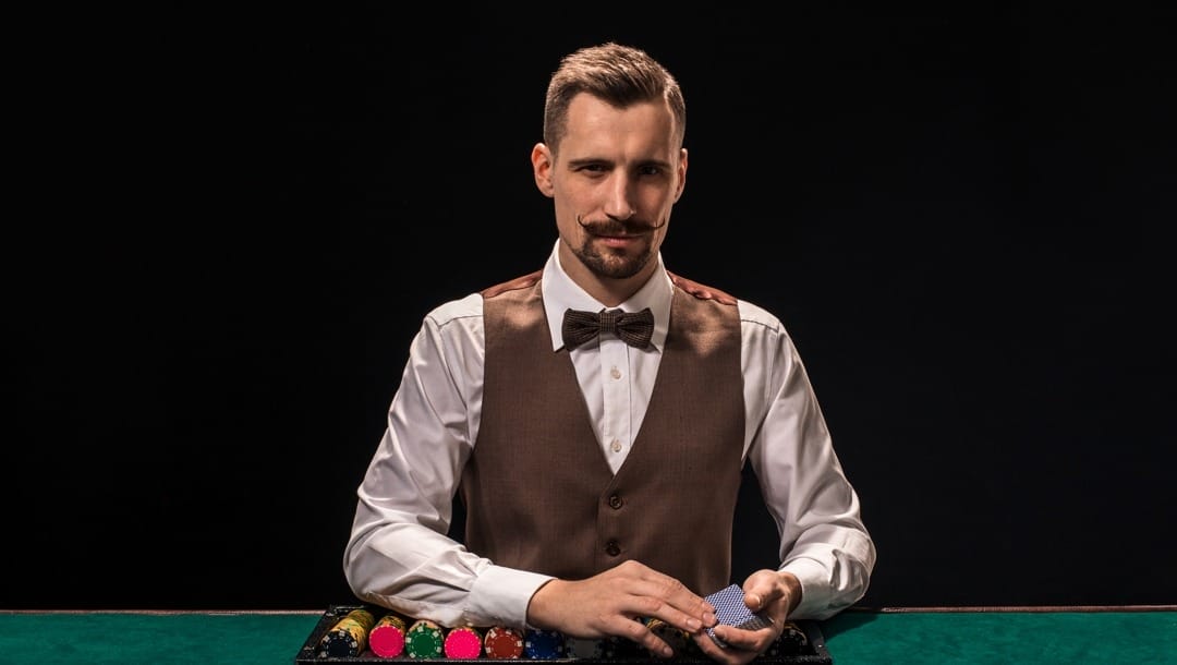 A well-dressed dealer with a beard and a curled mustache about to deal cards. There is a tray of poker chips on the table next to him.