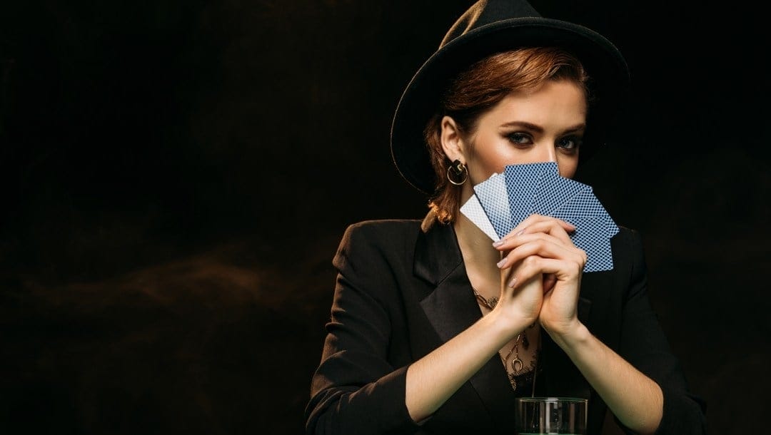 An elegantly dressed woman hides part of her face behind several playing cards.