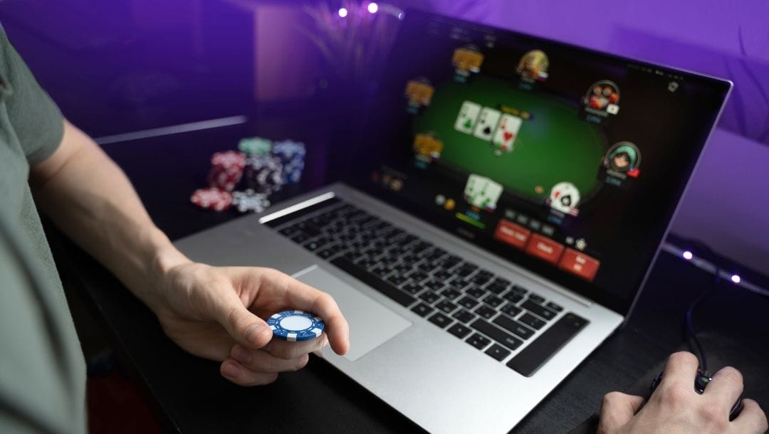 Man playing online poker on laptop, one hand on the mouse and the other holding a poker chip. Stacks of chips in the background.