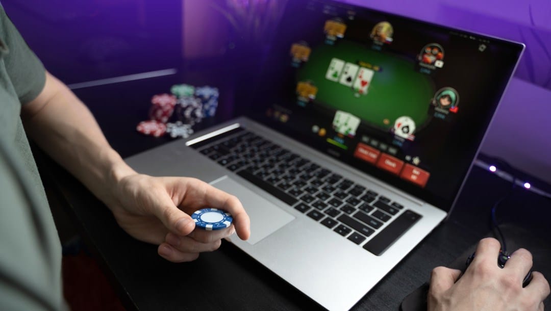 A man holding a casino chip while playing online poker on a laptop.