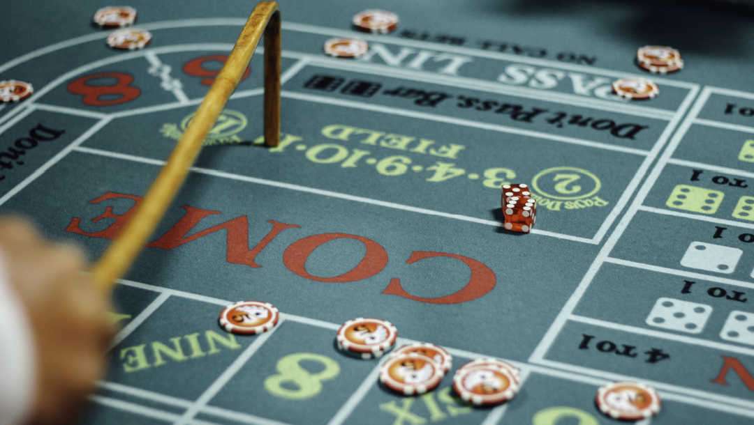 A casino dealer using a dice stick while two red dice sit on a craps table.