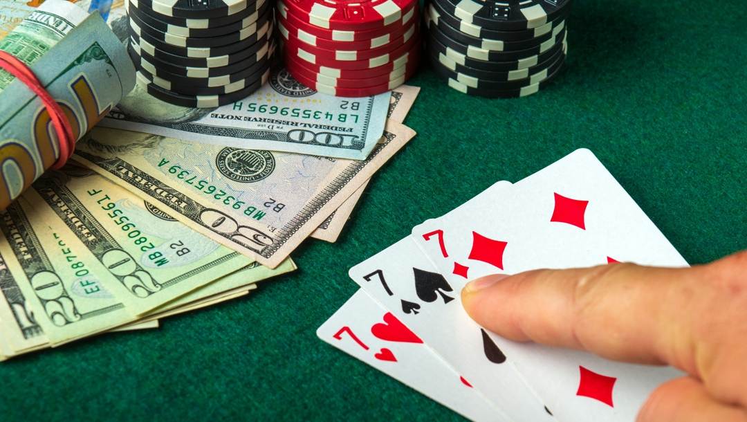 A player points at their three sevens poker hand with a roll of cash, loose dollar notes and poker chips in the background.