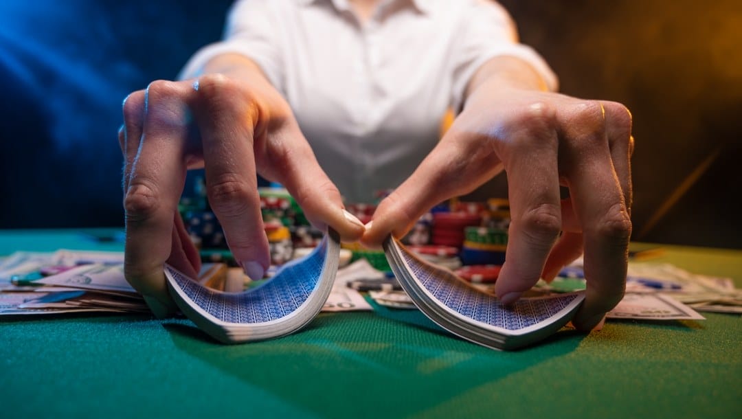 A close-up of a dealer’s hands shuffling cards. Cash and poker chips are scattered across the table behind their hands.