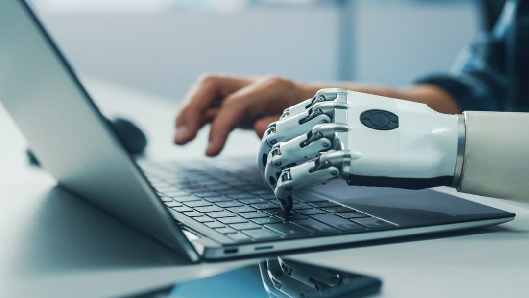 One human hand and one robotic hand typing on a laptop.