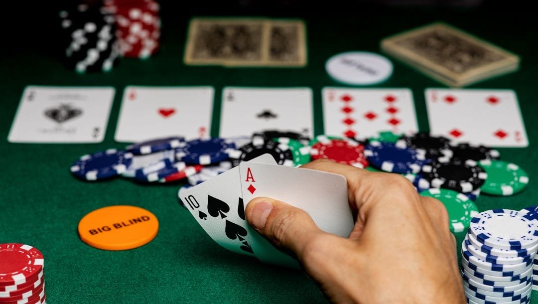 Player checking his hole cards of spade ten and diamond ace, in front of him on the poker table is a poker game comprising poker chips and playing cards post-flop