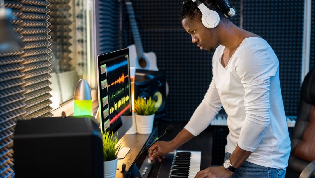 A young man standing in front of his computer and music keyboard mixing music.